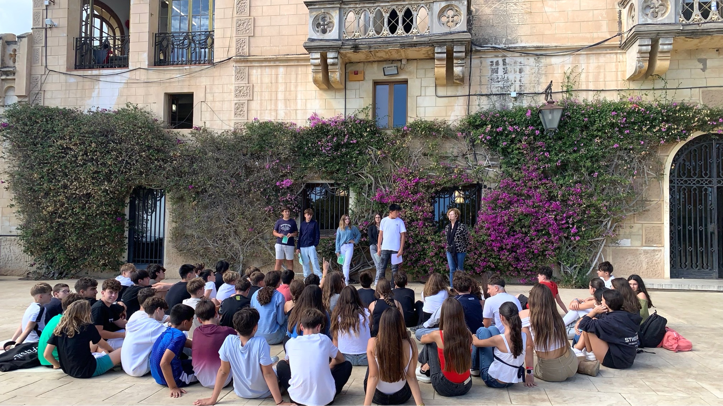 Los alumnos de una clase sentados en el suelo del patio escuchando a una docente