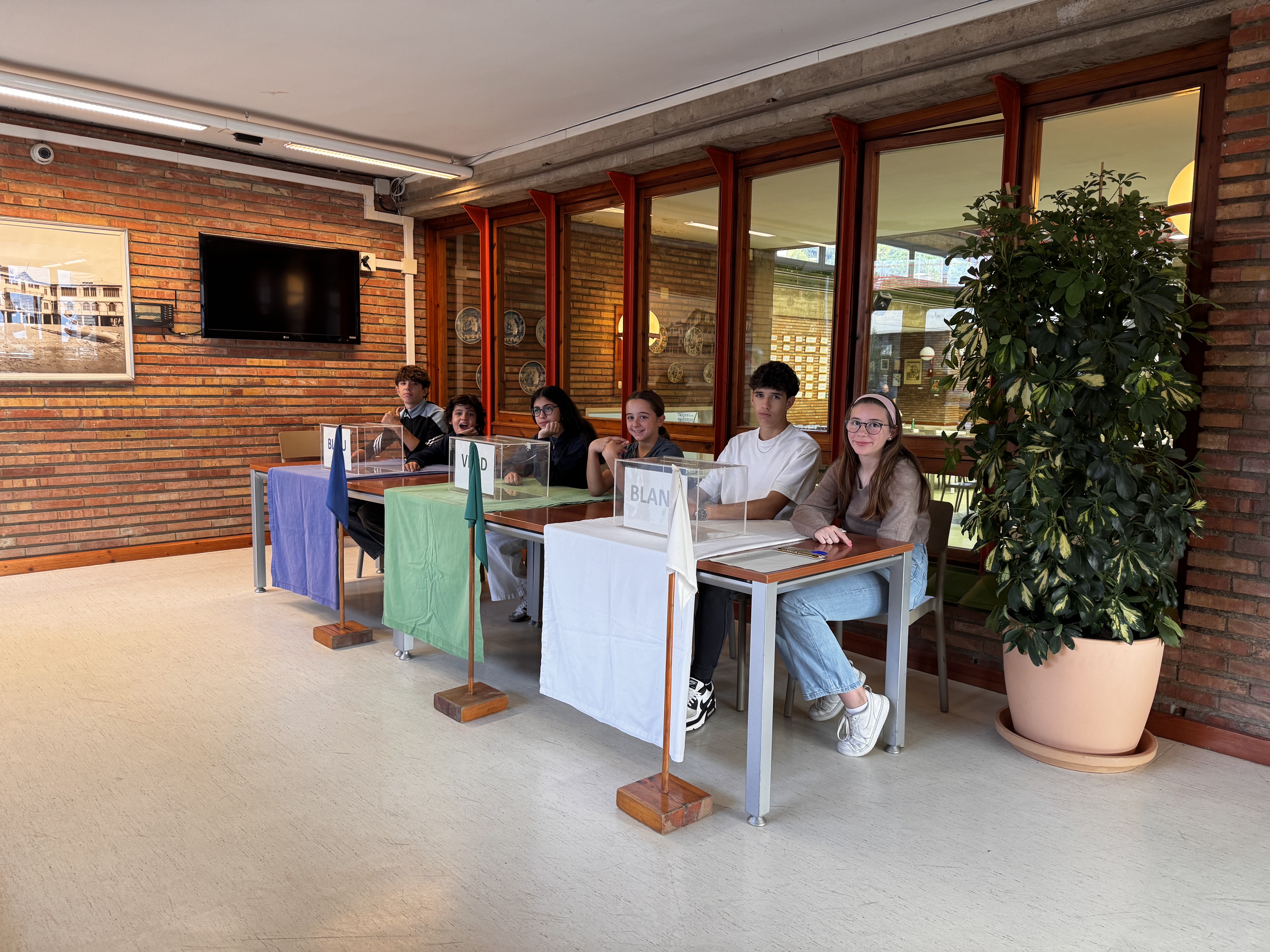 Cinco alumnos sentados en tres mesas electorales en el vestíbulo de la escuela